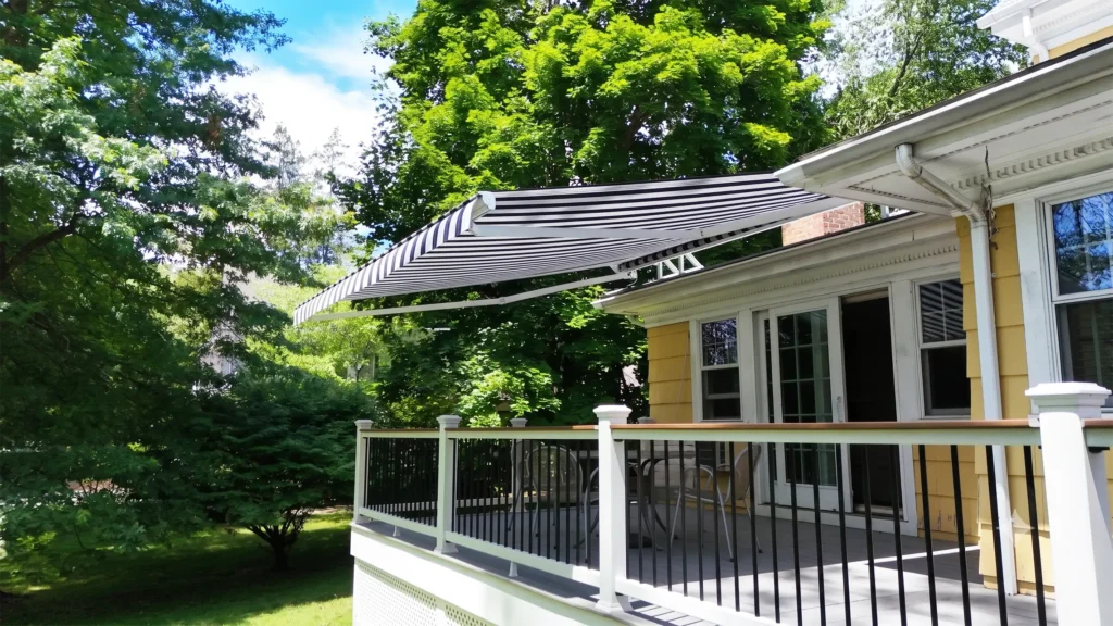 Striped retractable awning over a porch of a yellow house with white railings, in a sunny, green yard.