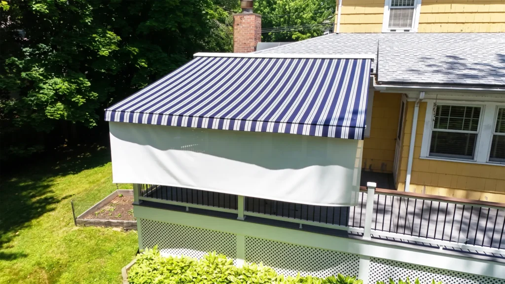 Blue and white striped awning extends over a balcony porch of a yellow house, with green trees in the background.